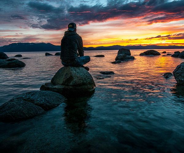 Silhouettes of two people sitting back-to-back in a meditative pose at sunset.